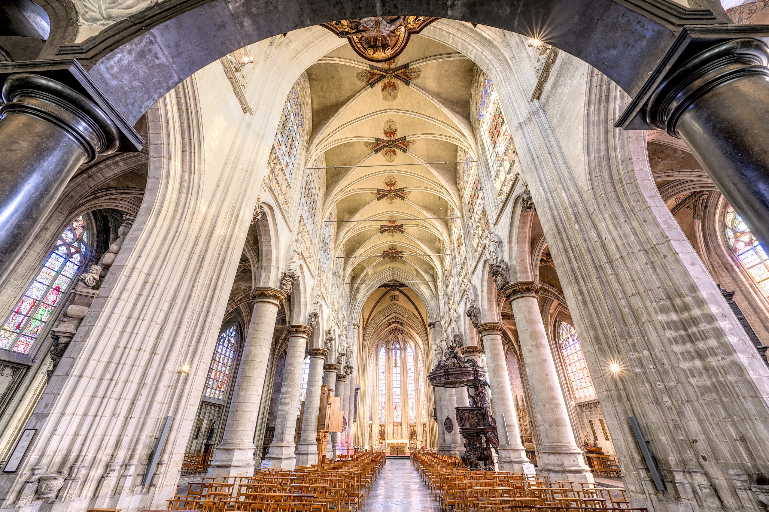 Binnenaanzicht van het schip van de kerk, verlicht met zuilen, een hoog plafond en gekleurde ramen.