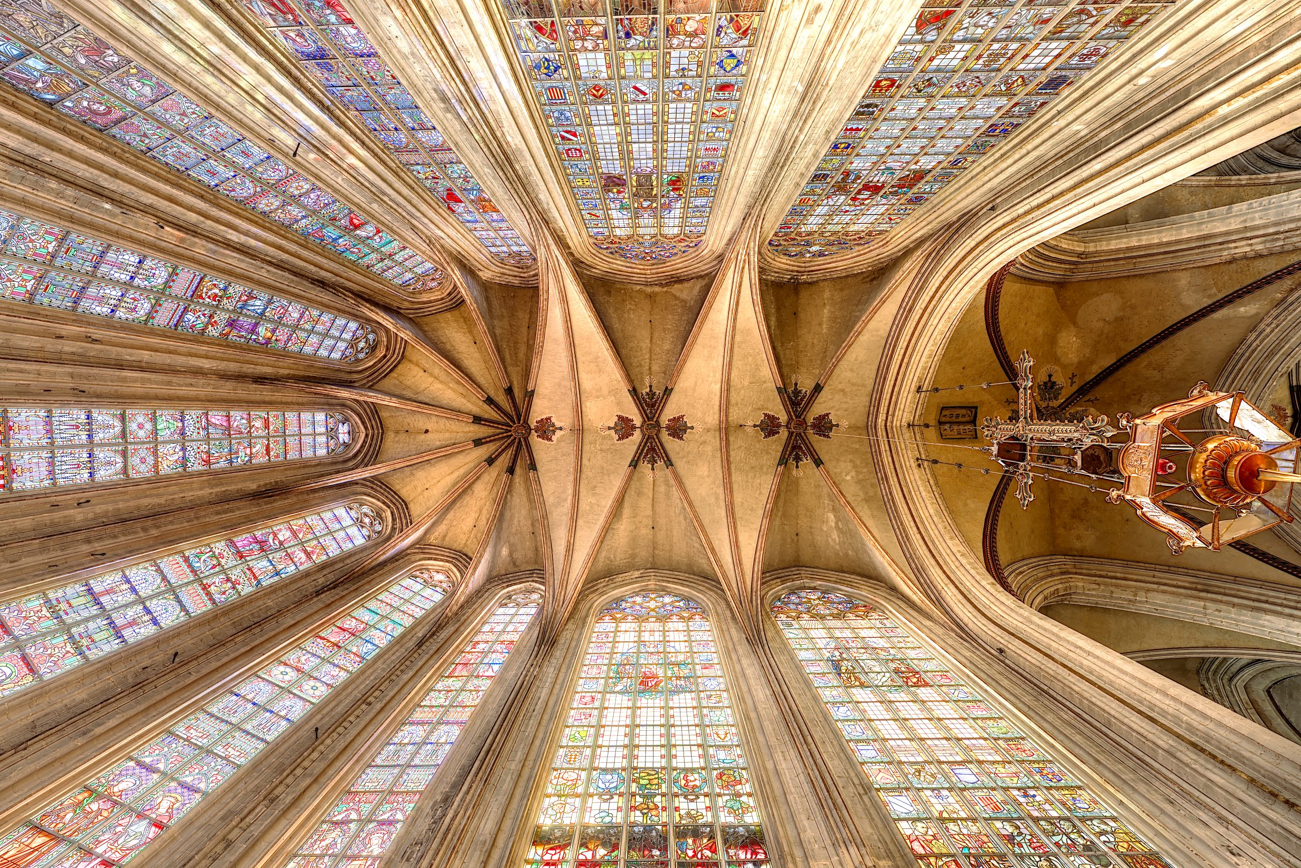 Zicht op het gewelfde plafond van de Notre-Dame kerk in Le Sablon, met gekleurde gebrandschilderde ramen en een grote hangende kroonluchter.